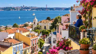 An older woman enjoys a balcony overlooking colorful hillside homes, blue water, and sailboats.