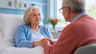 An older woman rests in a hospital bed while her spouse sits beside her and holds her hands.