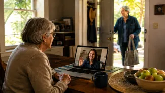 An older woman speaks with her distant daughter by video as a nearby helper arrives at the home.