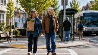 An older couple carrying groceries crosses a neighborhood street near a bus stop and community information board.