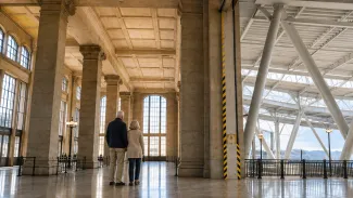 A retired couple stands where a historic terminal’s stone structure meets a separate modern support system.