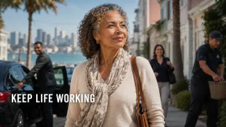 A biracial older woman walks confidently through a coastal neighborhood as transportation and household professionals provide support nearby.