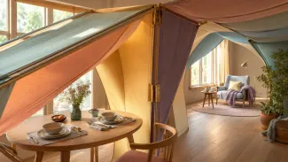 A soft fabric canopy connects a breakfast table set for two with a reading space for one, representing how the higher earner’s Social Security decision can affect both shared years and survivor years.