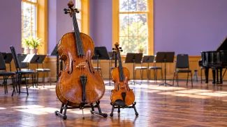 A well-used cello and violin stand in a sunlit community rehearsal room, ready for another session.