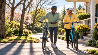 An older couple wearing helmets walks their bicycles along a sunlit path beside their home.