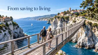 An older couple crosses a bridge above blue coastal waters toward a hillside town, beneath the words “From saving to living.”
