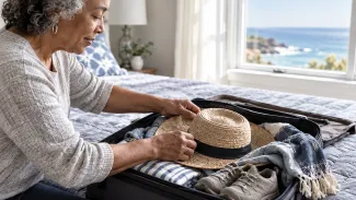 An older woman packs a sun hat and walking shoes into a suitcase beside a bright window overlooking the coast.