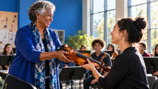 An older woman gives a violin to a music teacher in a sunlit community classroom.