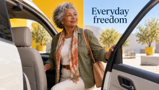 A confident older woman considers the comfortable passenger seat of a car in a bright outdoor plaza.