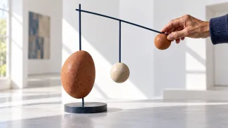 An older adult’s hand adjusts a small counterweight on a balanced sculpture of suspended stone forms.