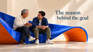 An older father and his adult son talk on a curved blue and orange bench beside the words “The reason behind the goal.”