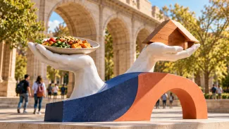 Two sculpted hands support a meal and a wooden house in a sunlit college courtyard.