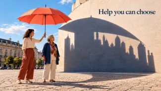 A younger woman holds a coral umbrella beside an older woman as an oversized architectural shadow falls across a wall.