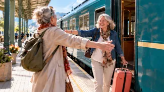 Two older friends reach to embrace beside a turquoise train, with a coral suitcase nearby.