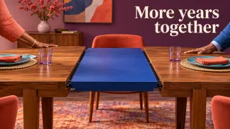Two people extend a wooden dining table with a blue center leaf beneath the words More years together.