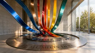 Colorful metal ribbons converge into one circular installation marked “It All Meets Here” in a sunlit gallery.
