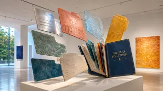 Colorful suspended document sculptures converge on an open volume labeled “The Year, Together” in a bright gallery.