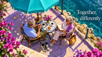 An older couple shares a seaside café table with a book and walking shoes beneath the words “Together, differently.”