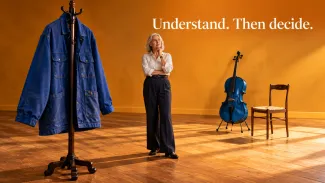 A woman stands between an oversized work jacket and a cello beneath the words “Understand. Then decide.”