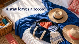 Two straw hats and an open travel bag rest on a sunlit vacation bed beneath the words “A stay leaves a record.”